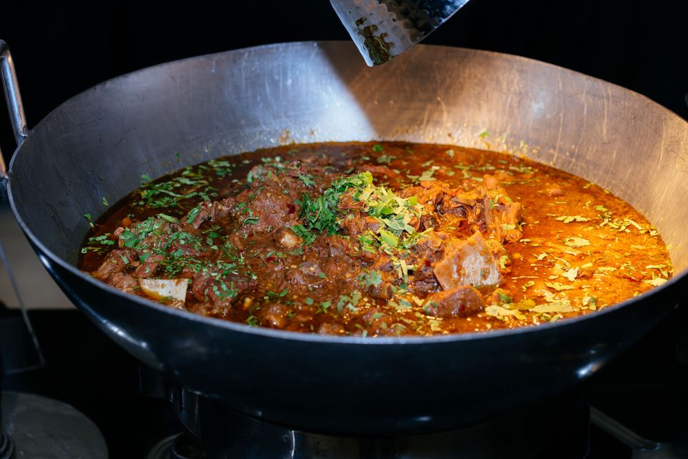 Rich meat curry with fresh green herbs simmering in a large black wok on a stove, served in a savory reddish-brown sauce.