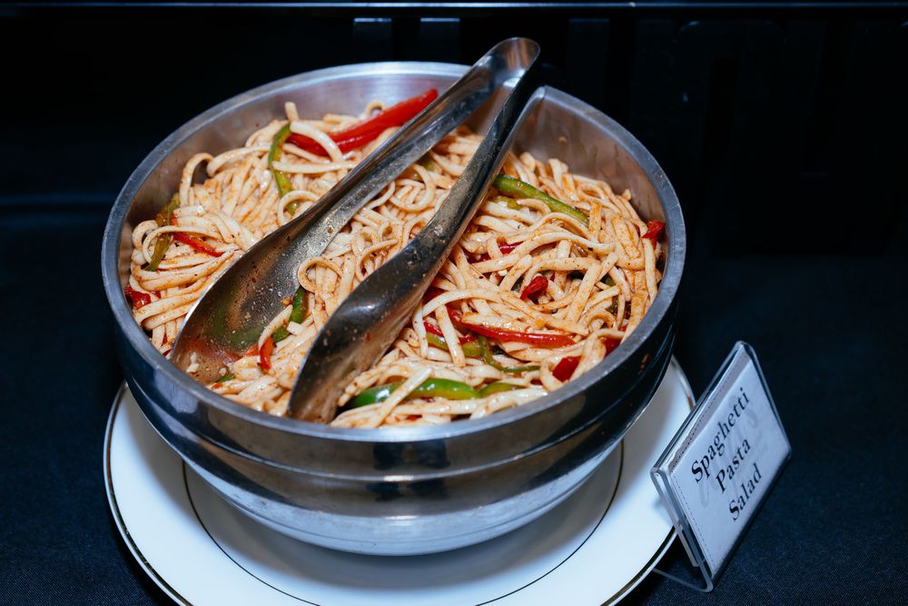 Spaghetti pasta salad with sliced red and green peppers served in a metal bowl with tongs and a labeled sign at a buffet.