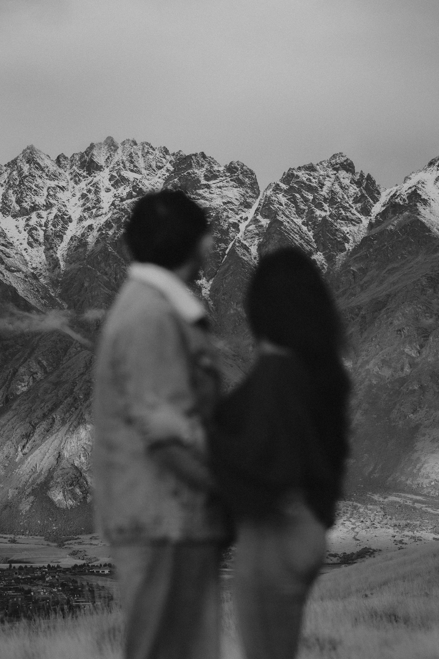 Blurred couple standing close together in the foreground, gazing at snow-capped rugged mountains in a moody black and white landscape.