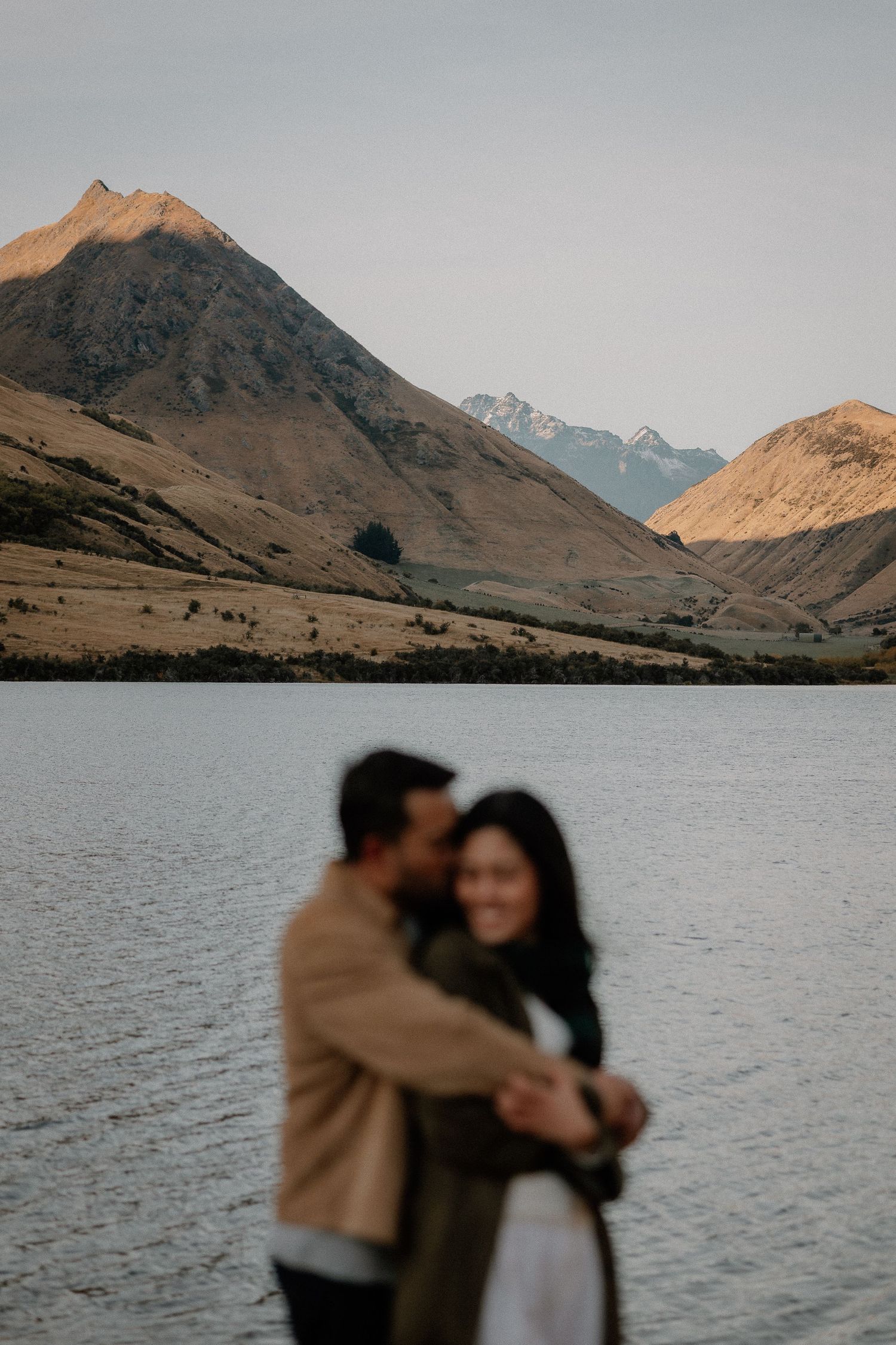 A couple embraces in soft focus beside a calm mountain lake, with golden hills and distant peaks rising behind them at sunset.