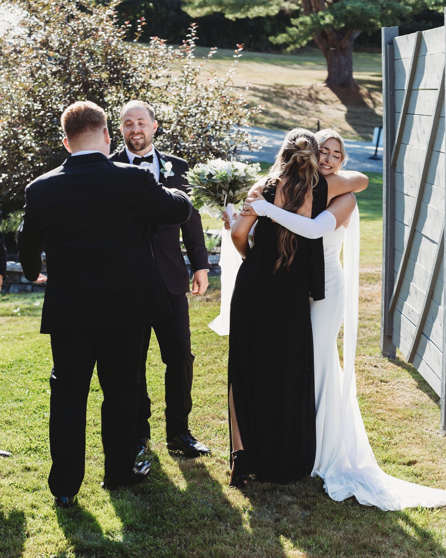 Bride hugging a guest outdoors while the groom smiles nearby.