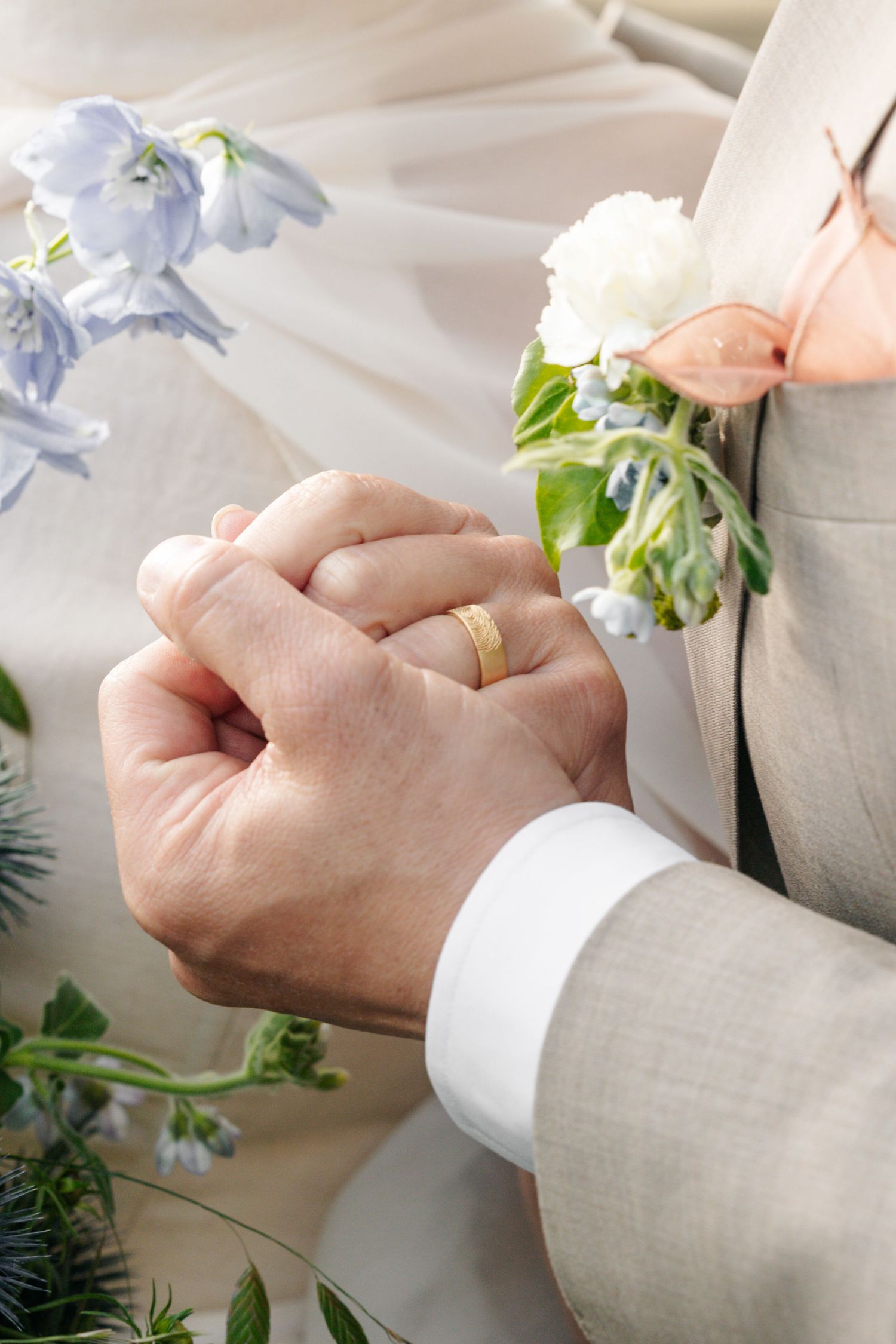 Groom in a tan suit with a white floral boutonniere clasps hands with his bride, a gold wedding band visible, beside blue wedding flowers.