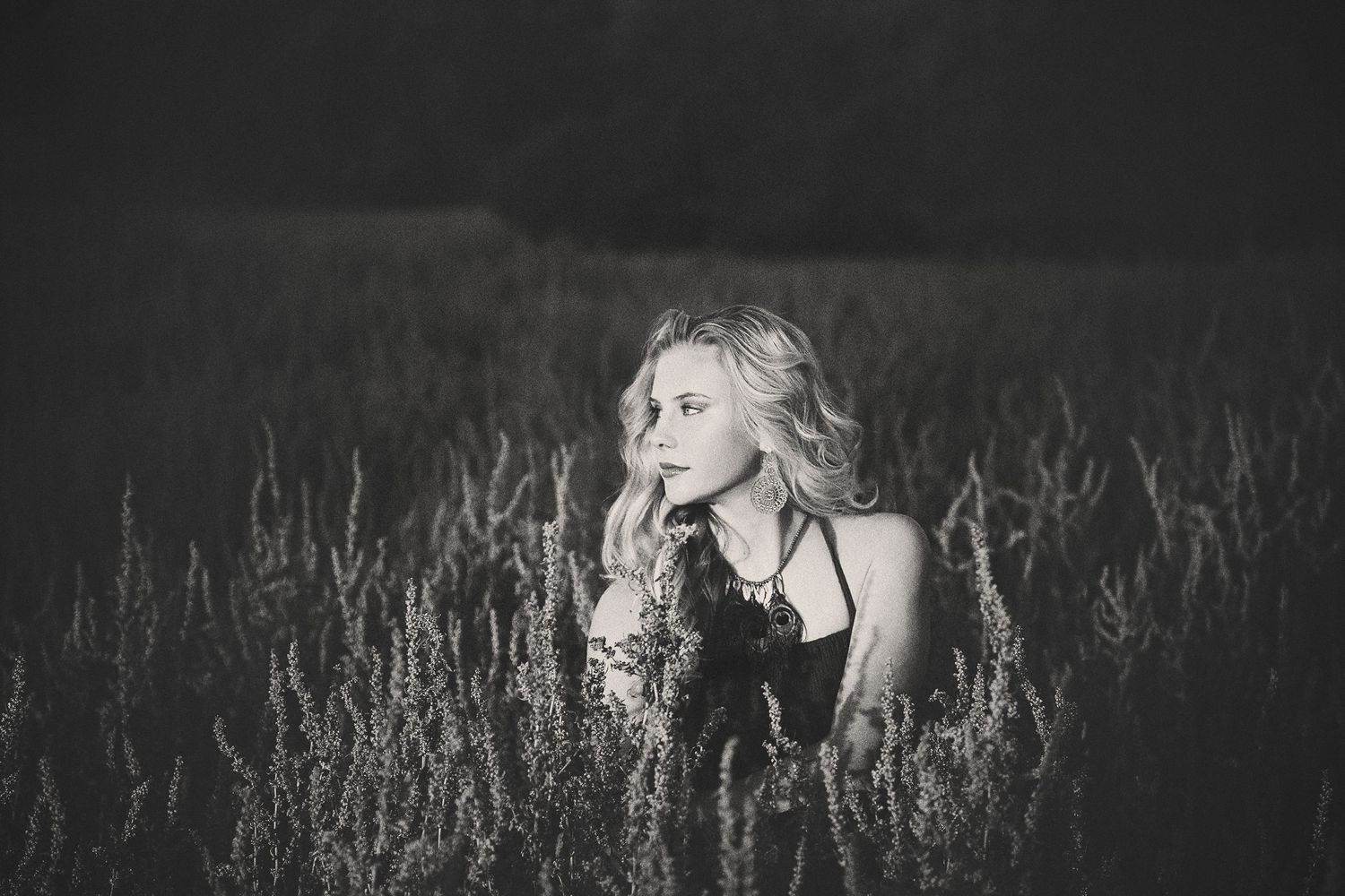 A black and white portrait of a young woman with wavy blonde hair sitting in tall wild grass and looking off to the side.