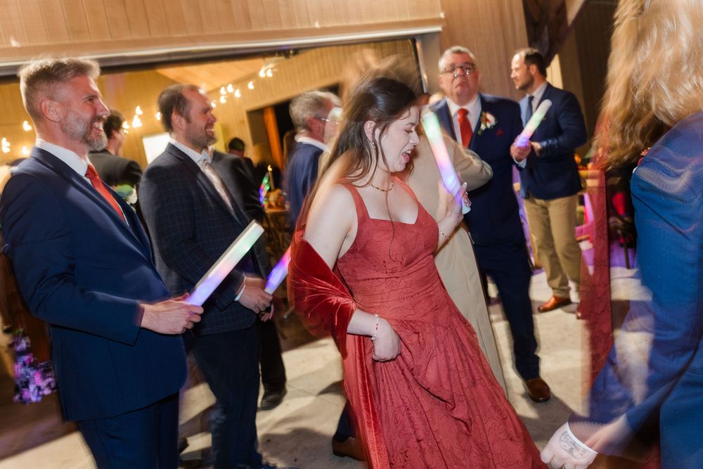 Woman in a red dress dances at an indoor evening reception while men in dark suits stand nearby holding glowing light sticks.