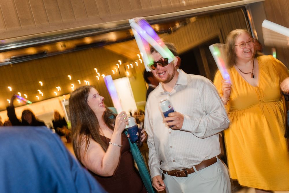 Smiling guests celebrate at an indoor party, wearing glow stick headbands and holding drinks under warm string lights.