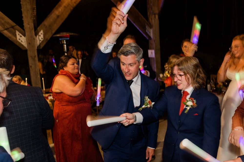 Two grooms in navy suits with boutonnieres dance joyfully at a wedding reception while guests wave glowing light sticks around them.