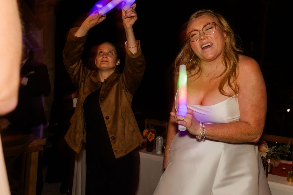 A smiling bride in a white strapless gown holds a glow stick while a woman in a brown jacket raises glow sticks behind her at a reception.