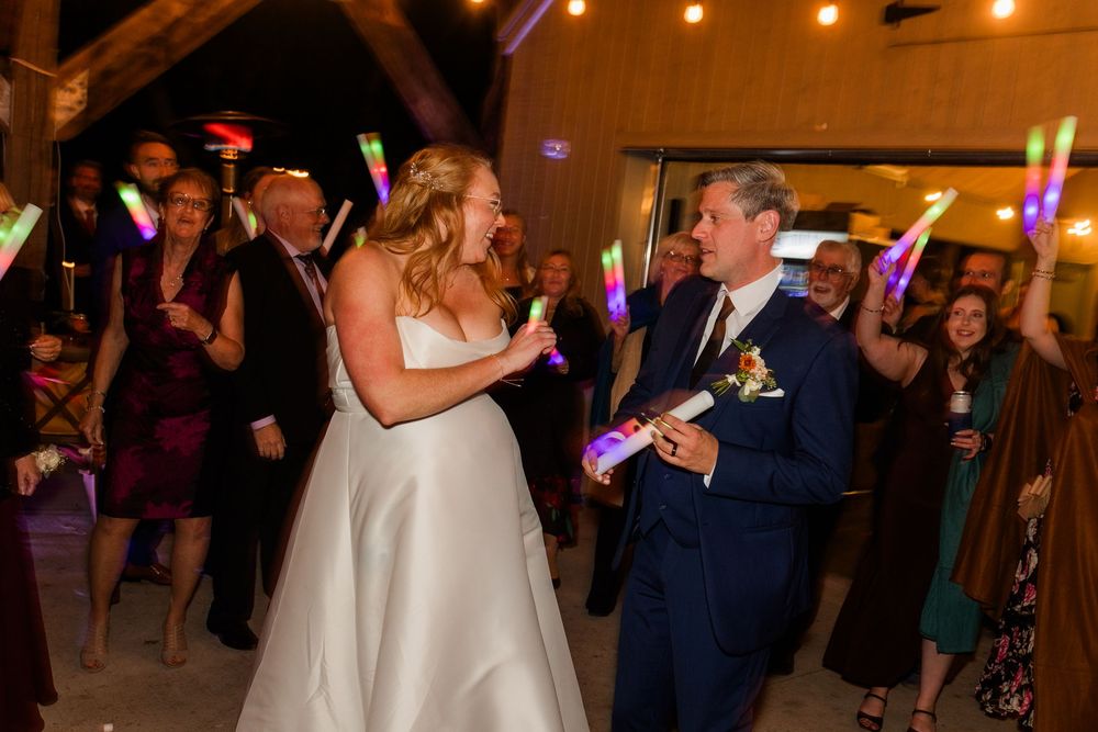 Bride in a white gown and groom in a navy suit smile while guests cheer with glowing light sticks during their wedding reception.