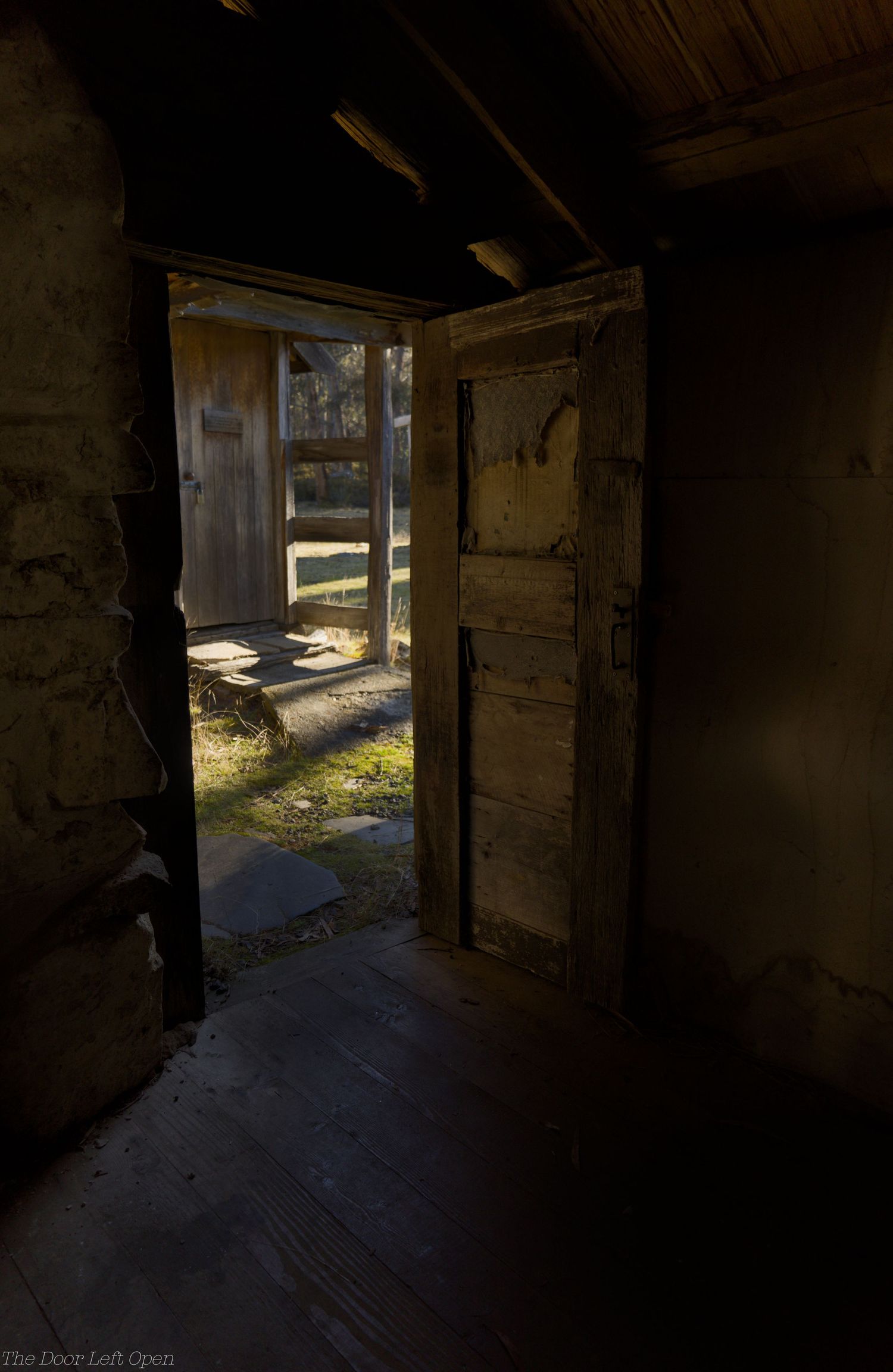 Dimly lit interior of an abandoned rustic cabin with a large stone fireplace, weathered wood plank ceiling, and light from a small window.