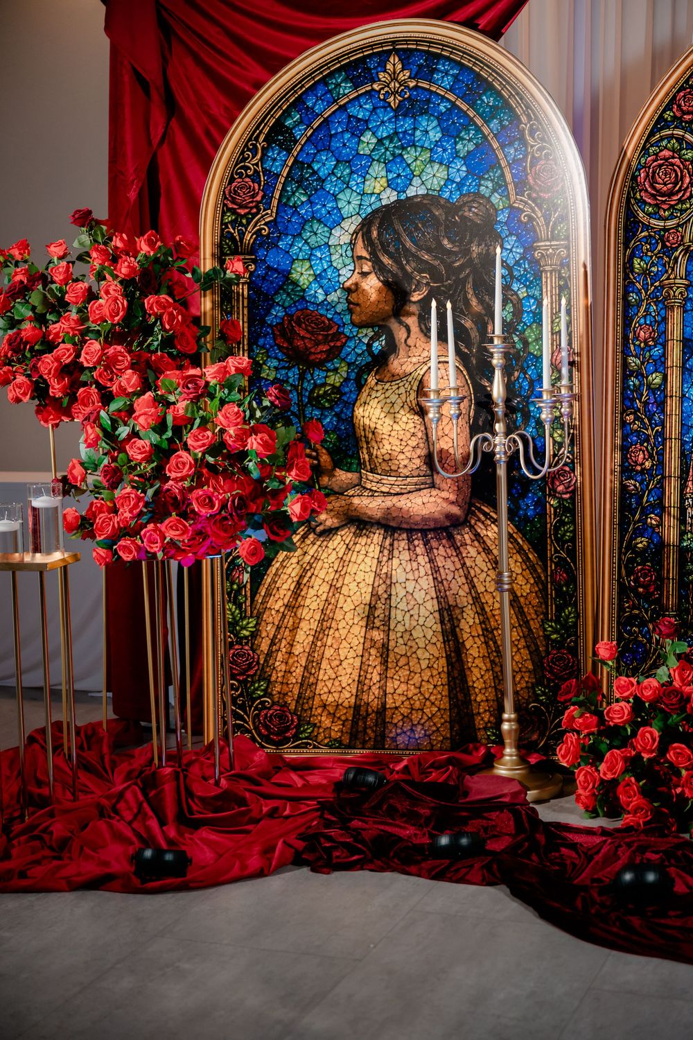 Stained glass style Belle display with golden gown, red roses, candelabra and draped red velvet at a fairytale themed event.