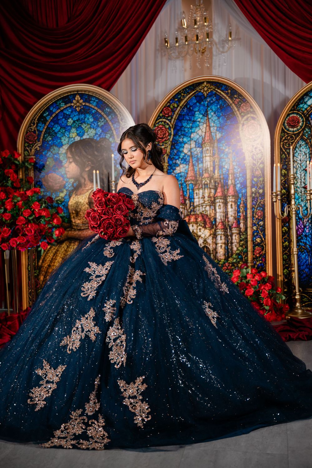 Young woman in a navy blue quinceañera ball gown with gold lace appliqués holding red roses before stained glass castle panels.