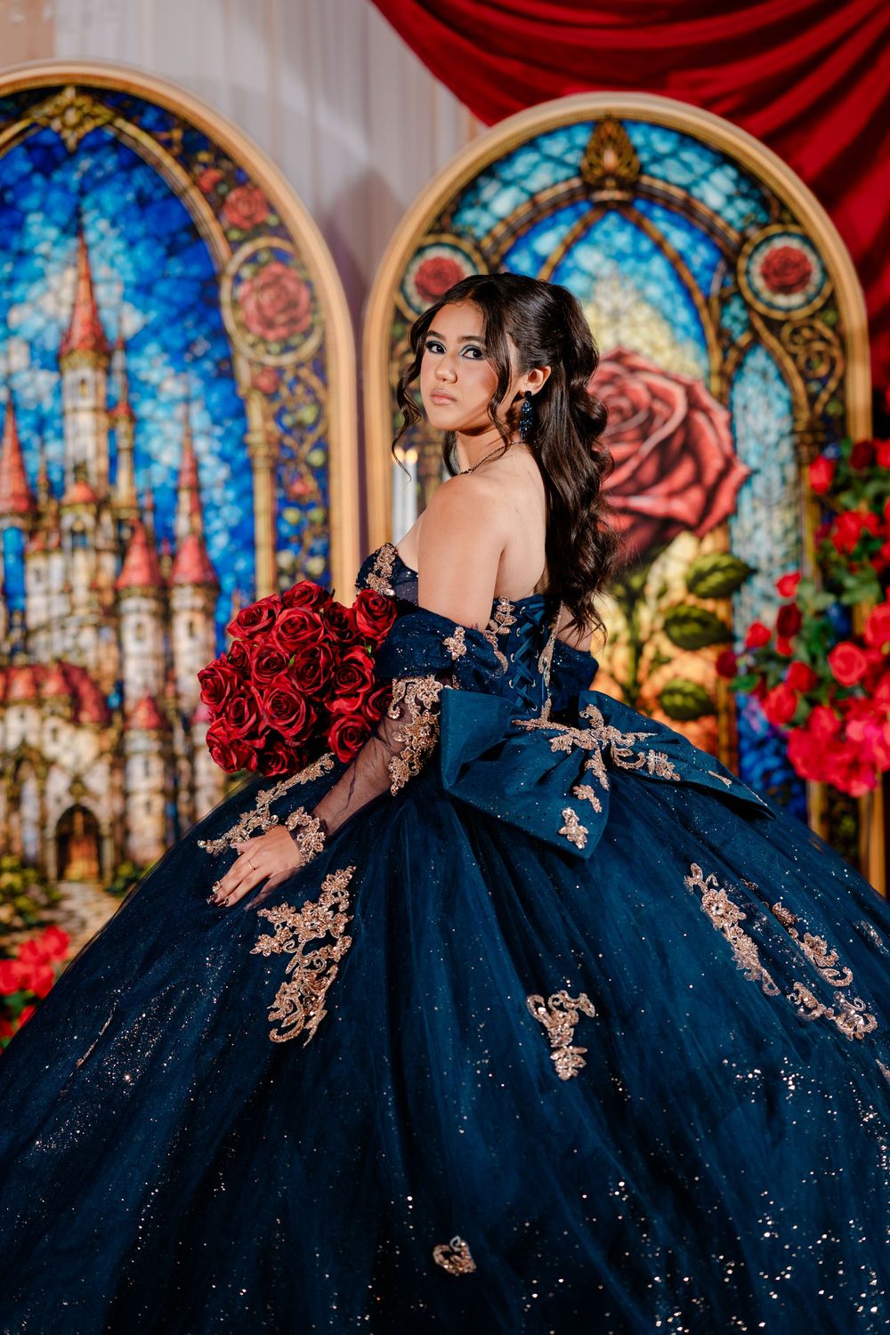 Young woman in a navy blue off-the-shoulder quinceañera ball gown with gold appliqués holds red roses before castle stained glass panels.