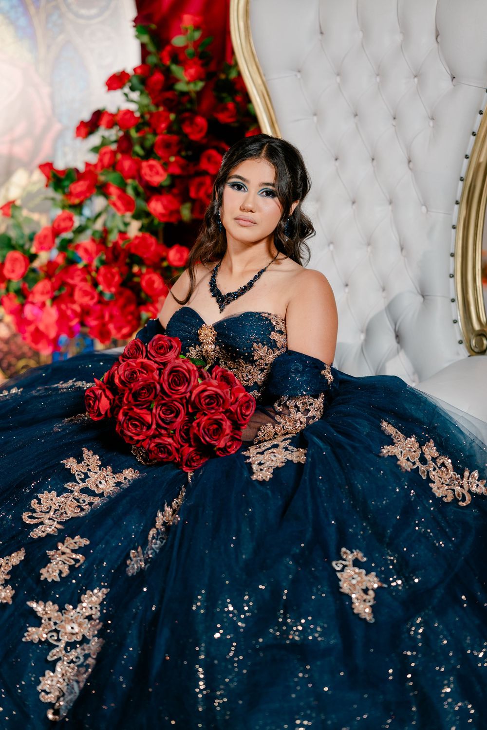 Young woman in a navy blue quinceañera ball gown with gold appliqués holds red roses while seated on a tufted white throne chair.