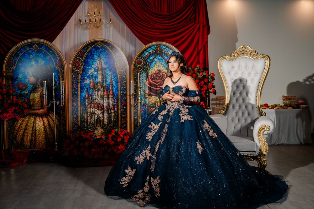 Young woman in an elegant navy blue quinceañera ballgown with gold floral embroidery poses beside a white throne chair and fairytale staine