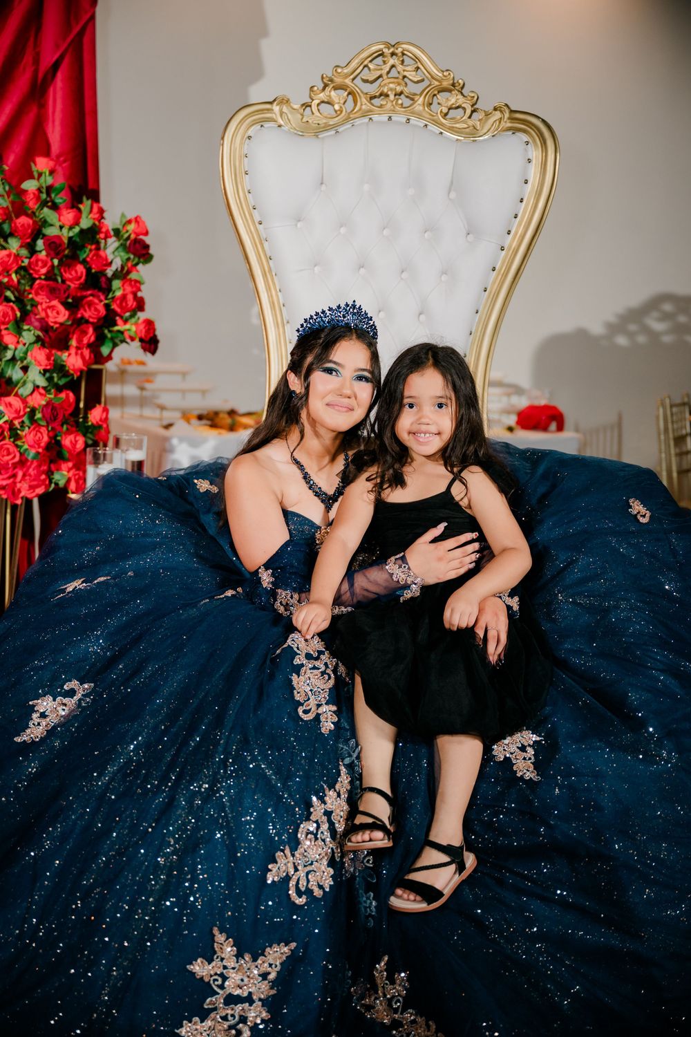 Young woman in a navy blue quinceañera gown and tiara sits on a white throne chair holding a little girl in black beside red roses.