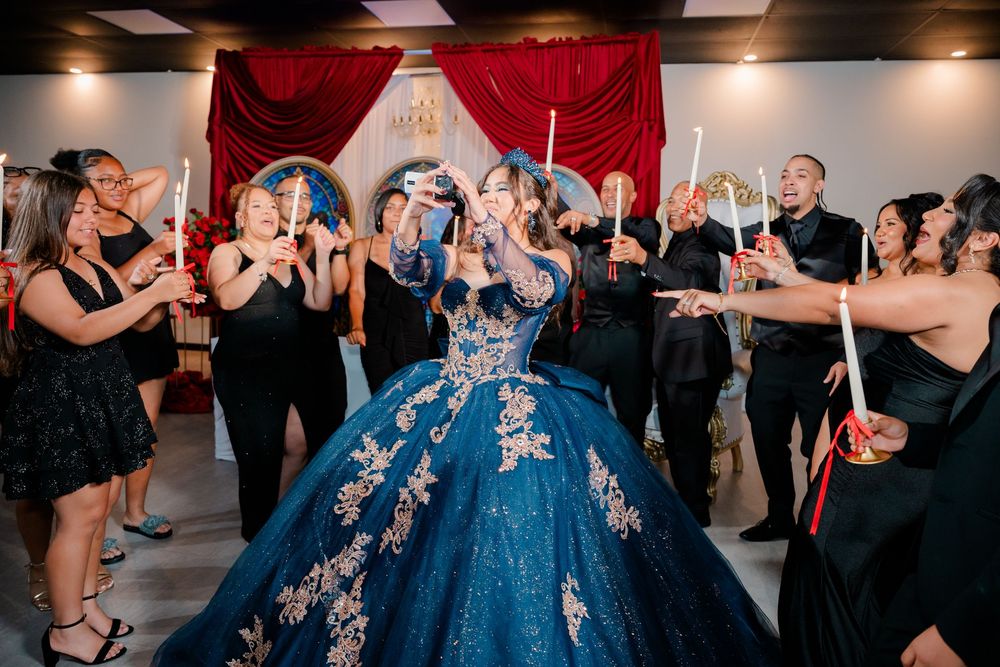 Quinceañera in a sparkling blue ball gown stands at the center of a ceremony as family and guests surround her holding lit candles.