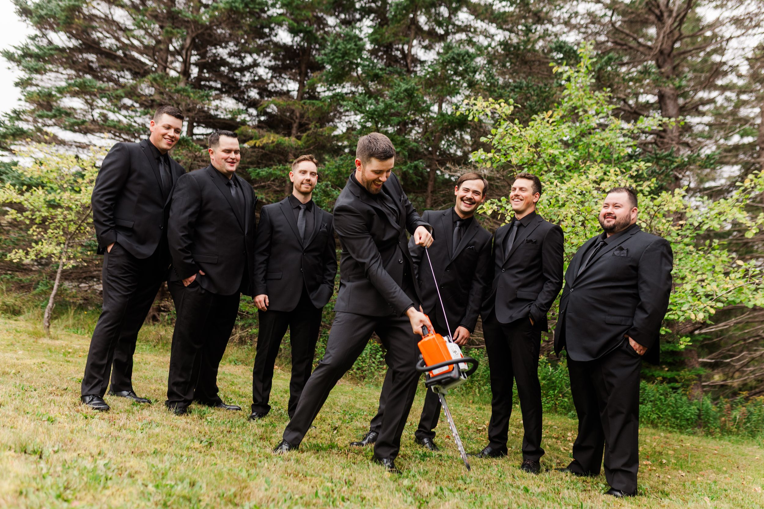 Six groomsmen in black suits laugh on a grassy lawn as one man bends over to start an orange gas-powered auger near evergreen trees.