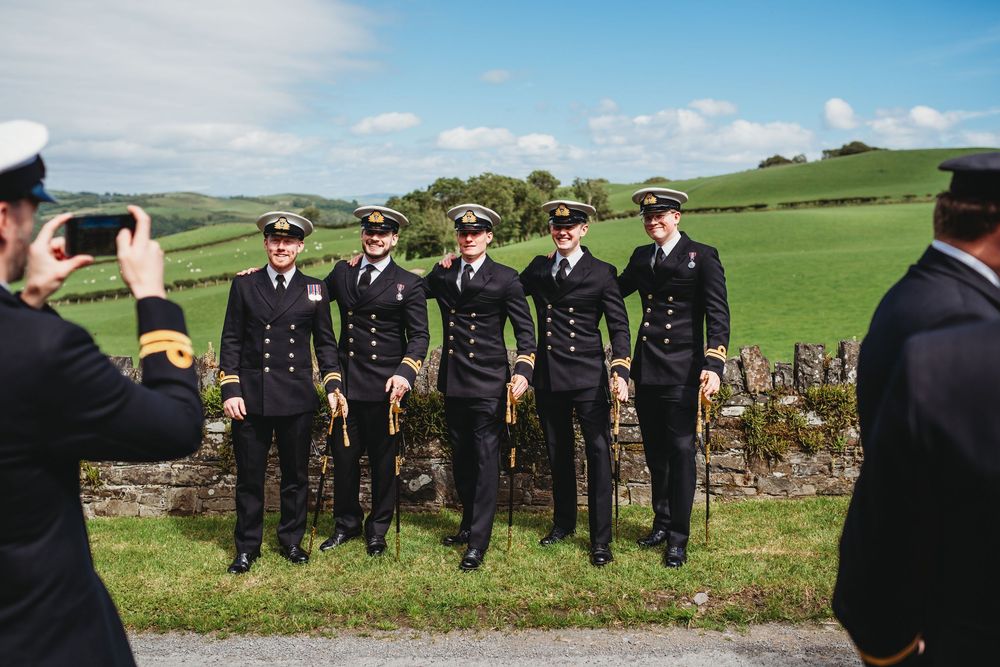 Four naval officers in dress uniforms pose outdoors near a stone wall with green hills behind them.