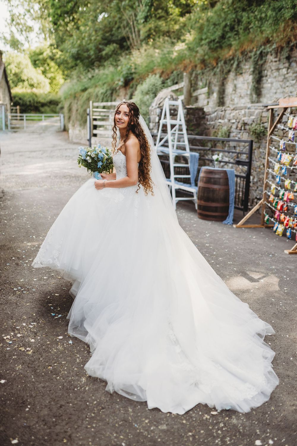 Smiling bride in a voluminous white ballgown with long train holding a blue bouquet outdoors.