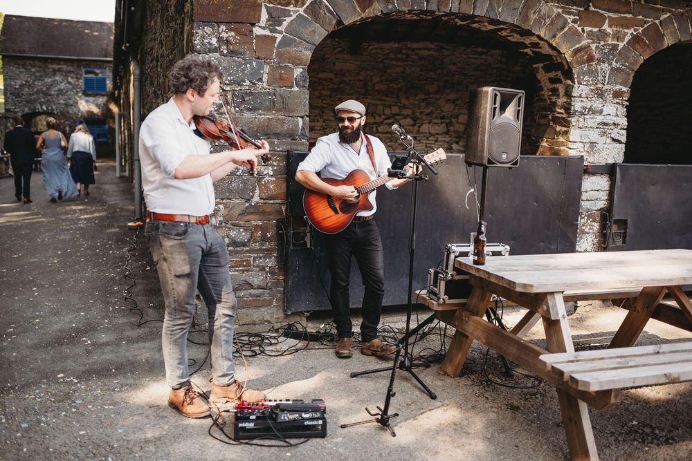 Two musicians perform outdoors, one playing violin and one playing acoustic guitar, against a rustic stone wall backdrop.
