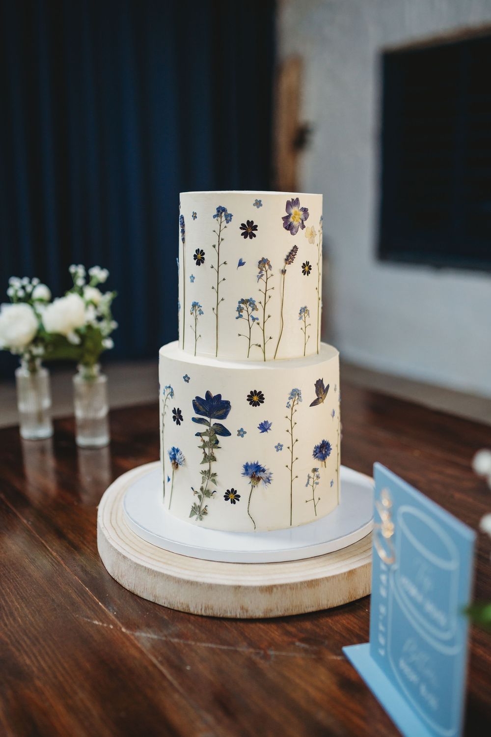 Two-tier white wedding cake decorated with pressed blue wildflowers and botanical illustrations on a wooden cake stand.