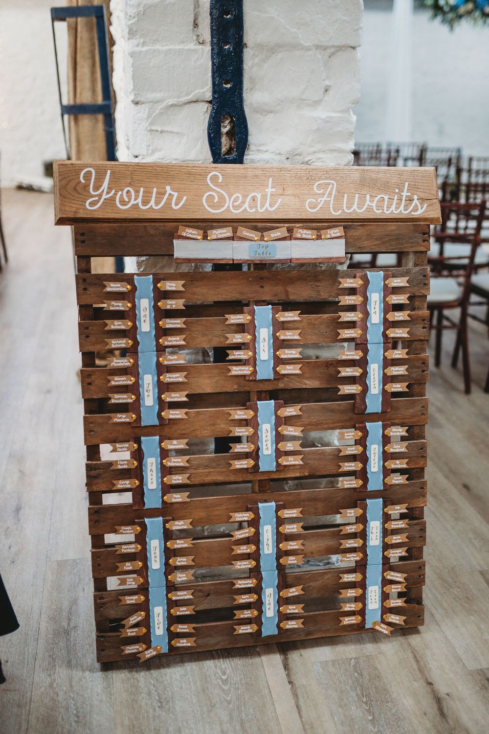Rustic wooden pallet wedding seating chart with blue ribbon name cards and "Your Seat Awaits" sign.