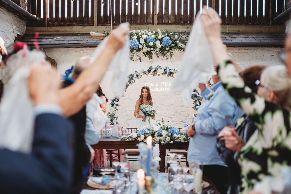 Elegant barn wedding reception with guests raising napkins as bride stands before floral arch backdrop with blue and white flowers.