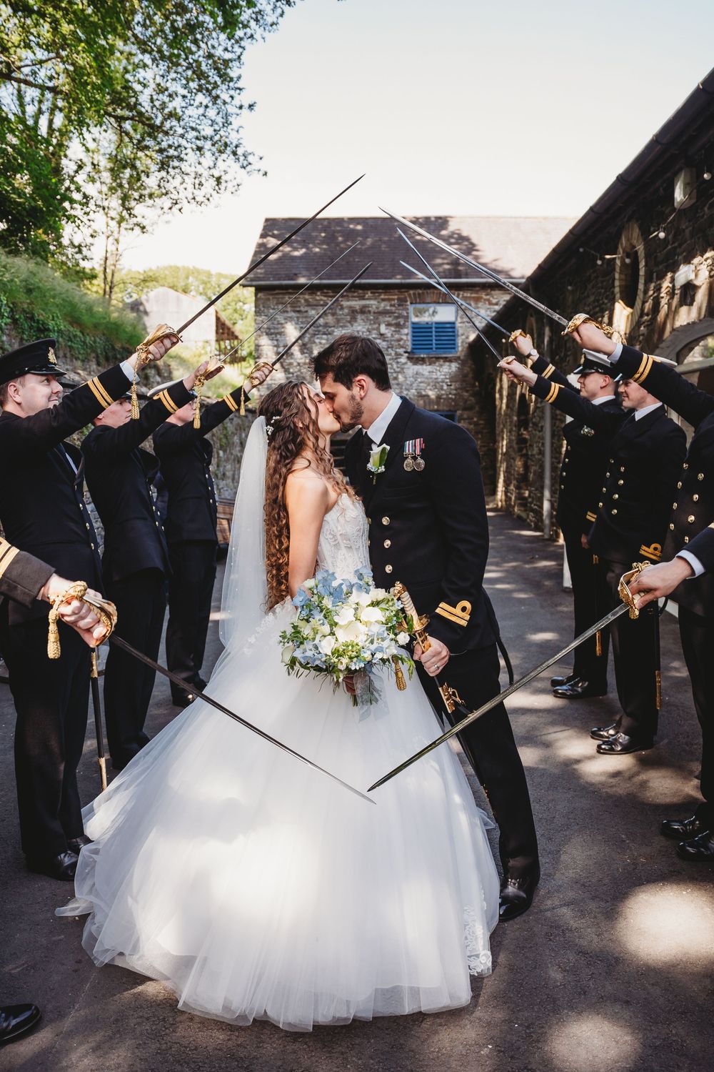 Naval officer groom kisses bride in white ballgown during military sword arch wedding ceremony outdoors.
