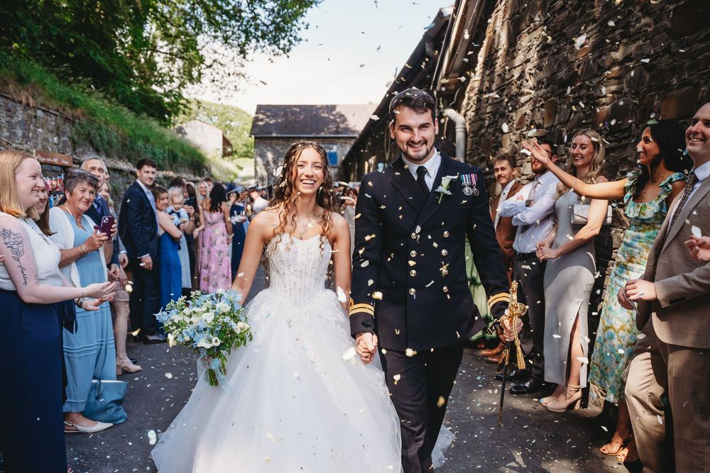 Bride and groom walk through confetti shower after wedding ceremony, surrounded by cheering guests outdoors.