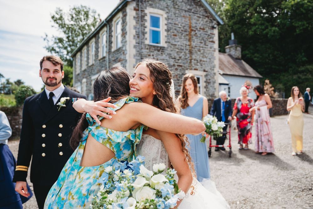Bride hugging a guest in a floral dress outside a stone building at a wedding celebration.