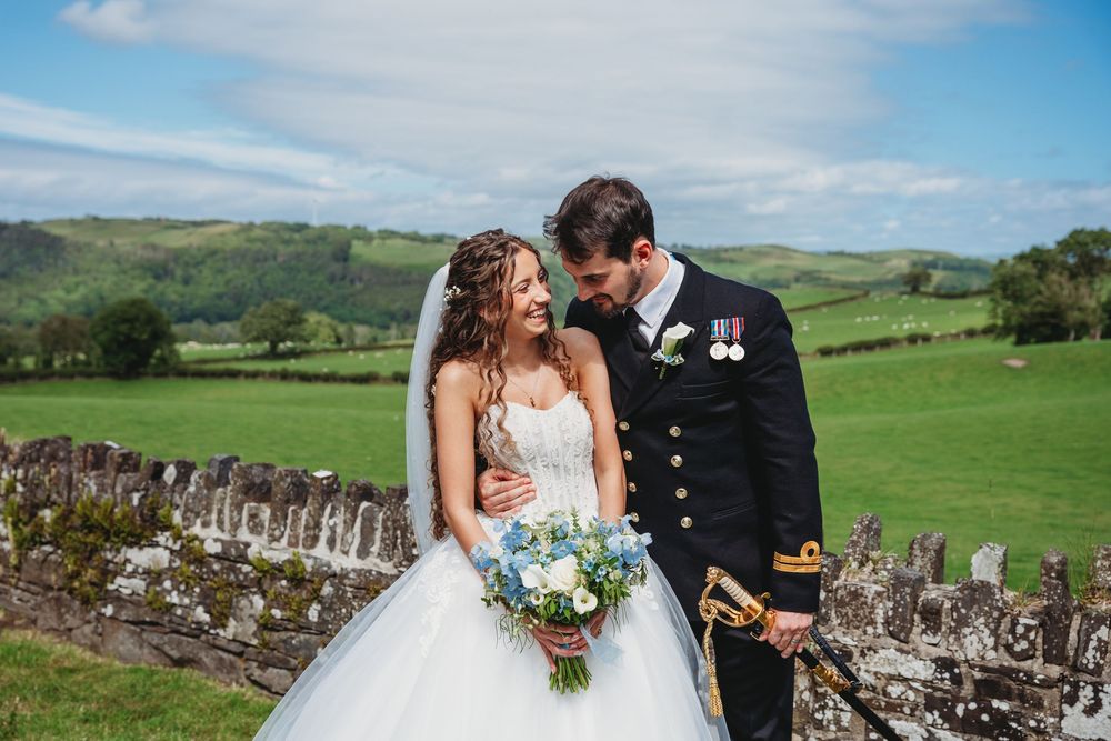 Bride and groom share a romantic moment outdoors, with rolling green hills and a stone wall backdrop on their wedding day.