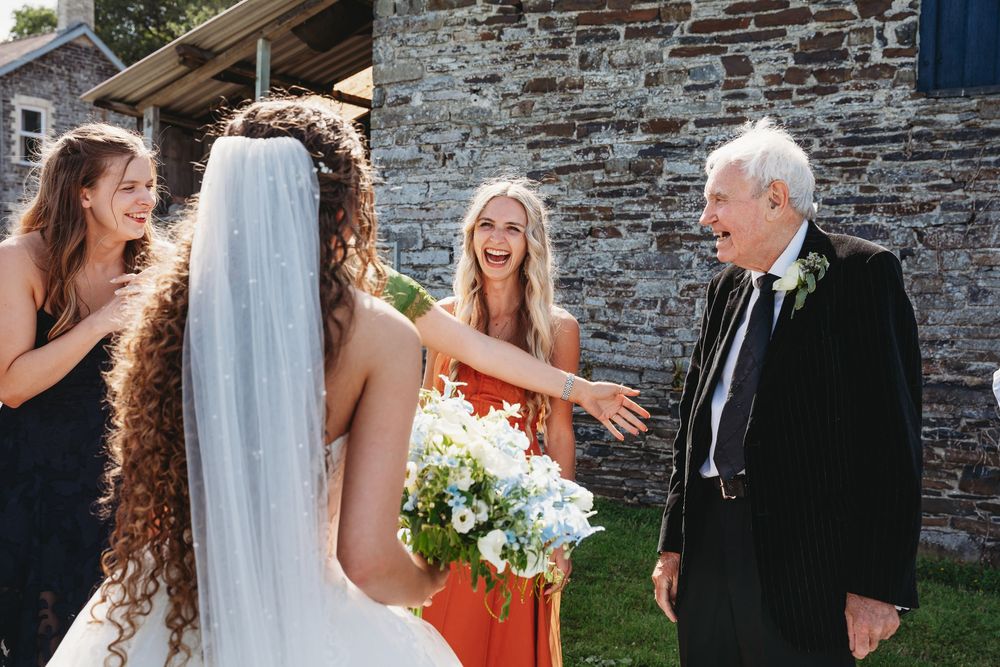 Bride in white gown and veil holds bouquet while laughing with bridesmaids and elderly man at outdoor wedding.