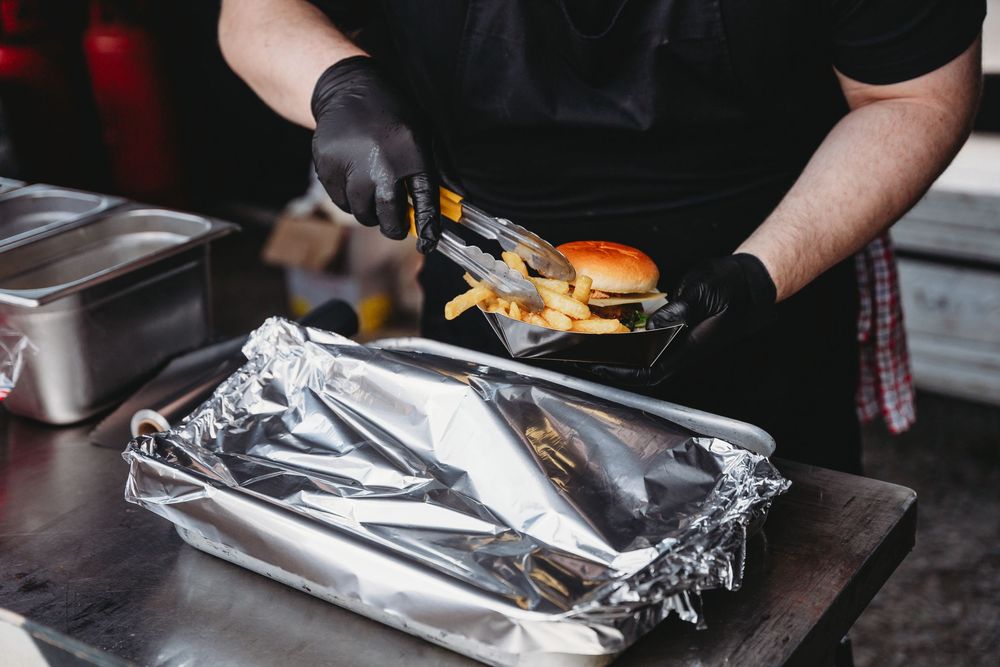 Chef in black gloves using tongs to place fries beside a burger on aluminum foil in a commercial kitchen.