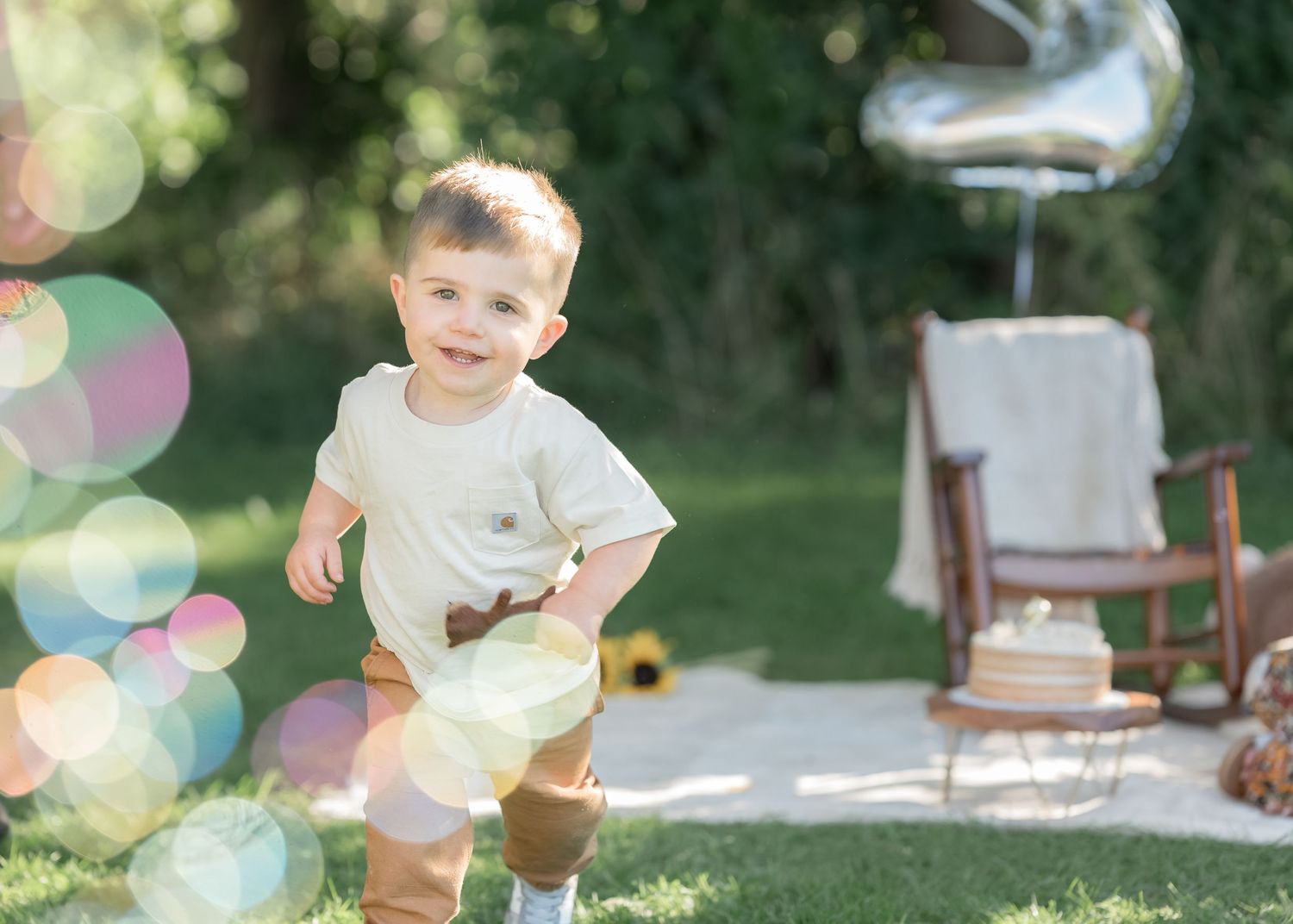 Smiling toddler boy in white shirt running outdoors at a first birthday party with balloons and cake.