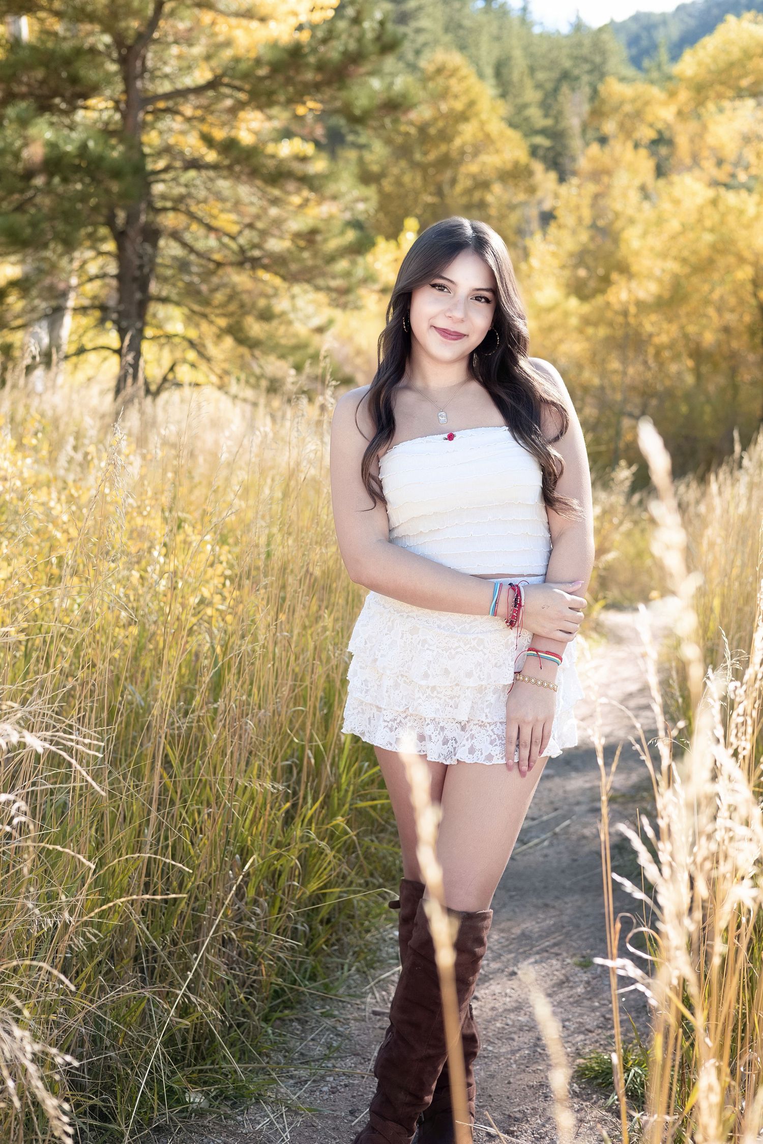 Young woman in white top, lace shorts, and brown knee-high boots posing on a path surrounded by tall golden grass and trees.