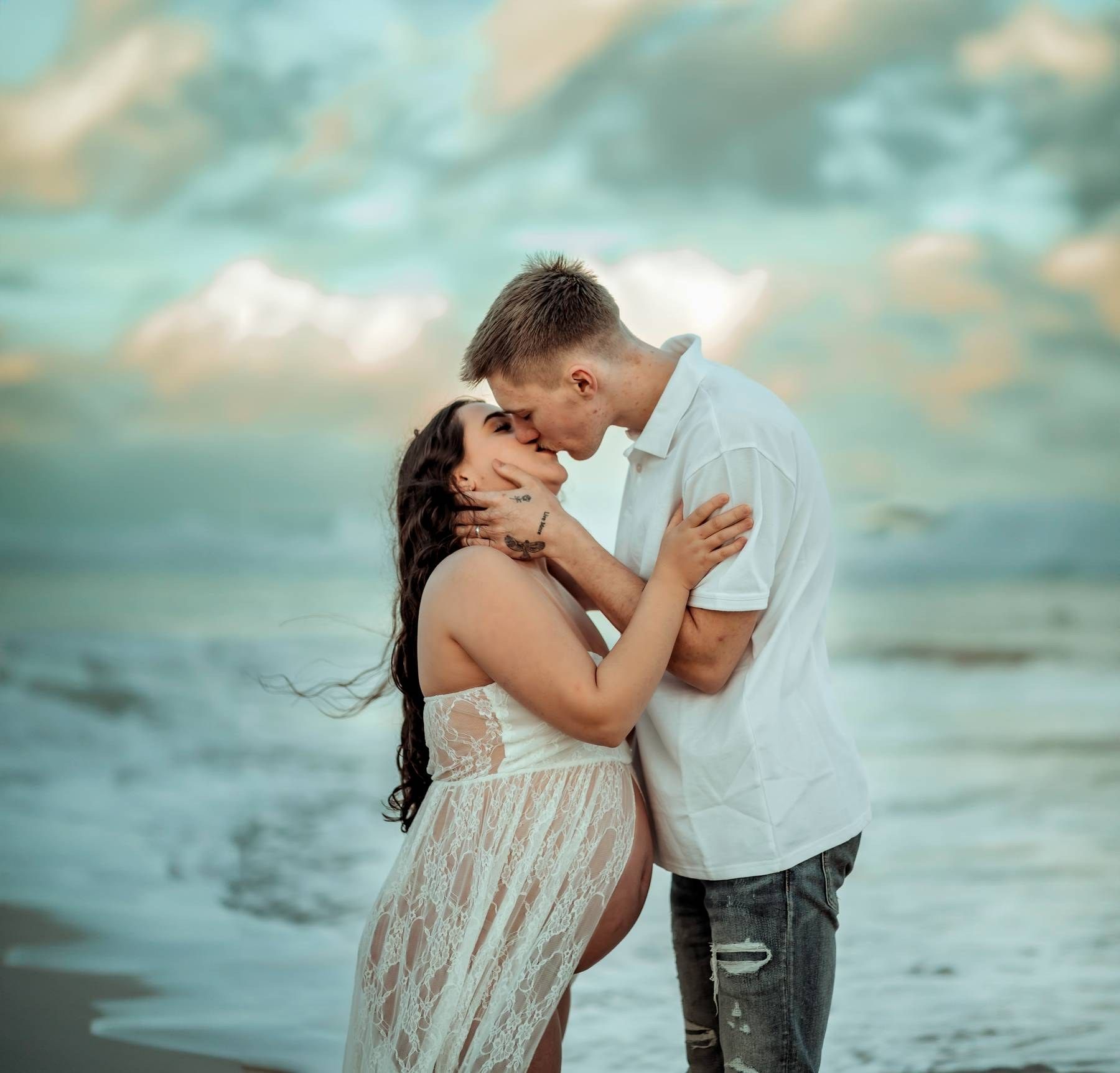 A romantic couple embraces and kisses on a beach at sunset, woman in white lace dress, man in white shirt.