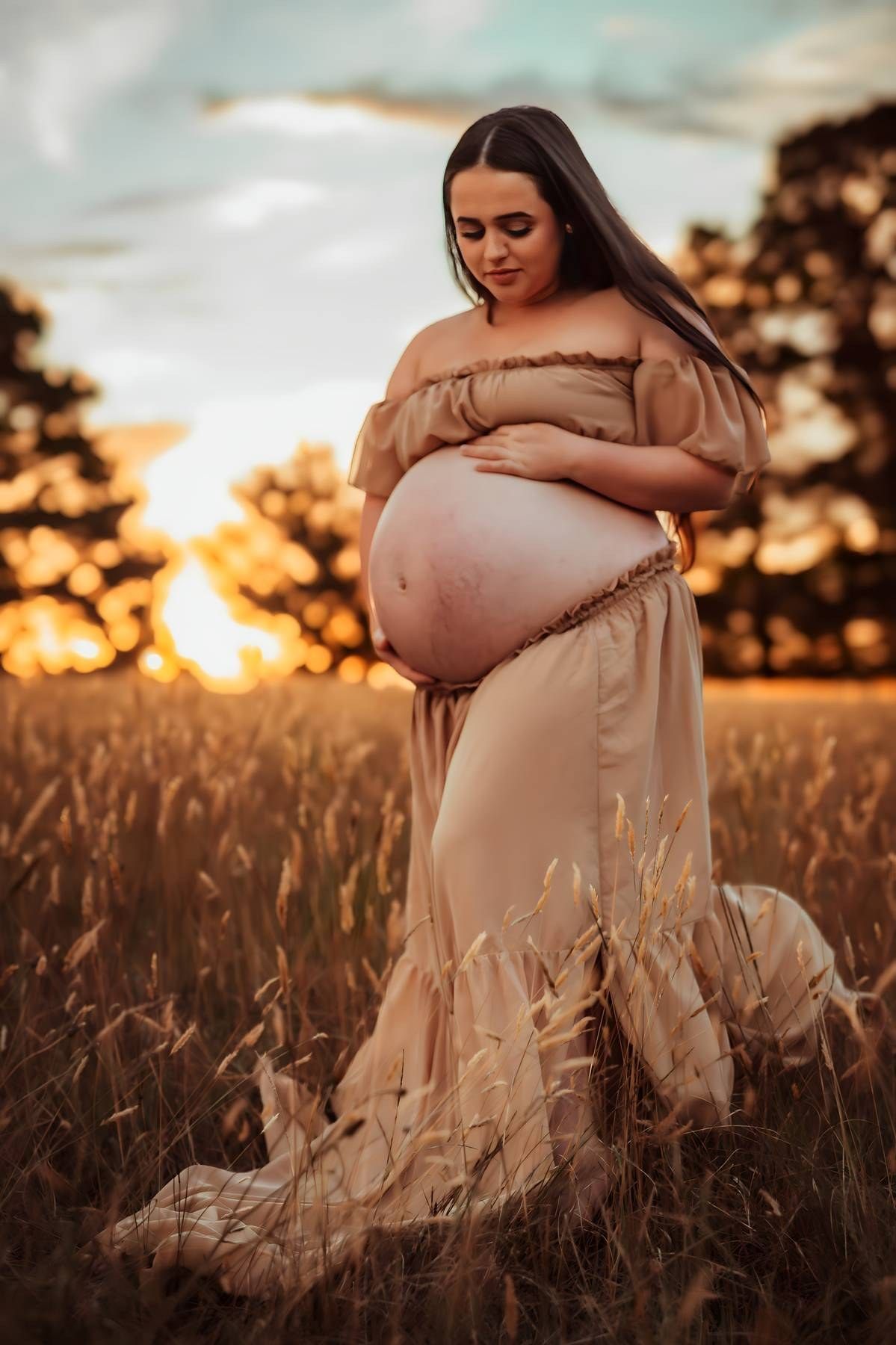 Pregnant woman in beige off-shoulder gown cradling belly in golden wheat field at sunset.