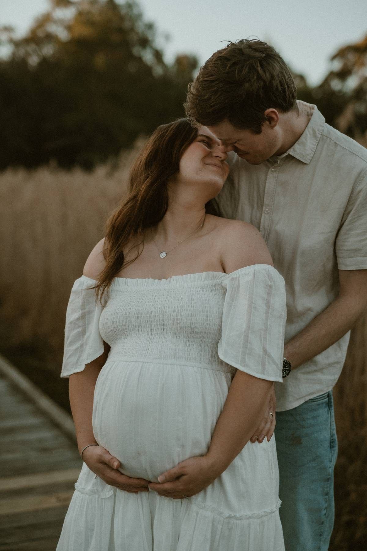 Pregnant couple sharing a tender moment on a wooden boardwalk, woman in white off-shoulder dress cradling her baby bump.
