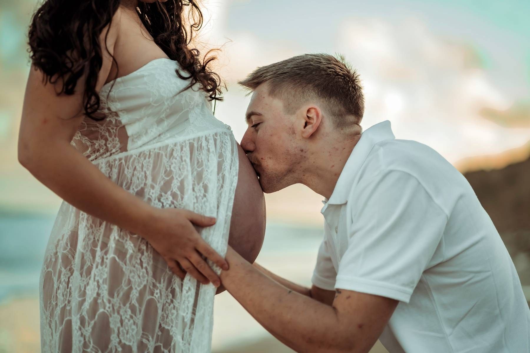 Man kissing pregnant woman's belly on beach at sunset, maternity photo shoot with couple in white outfits.