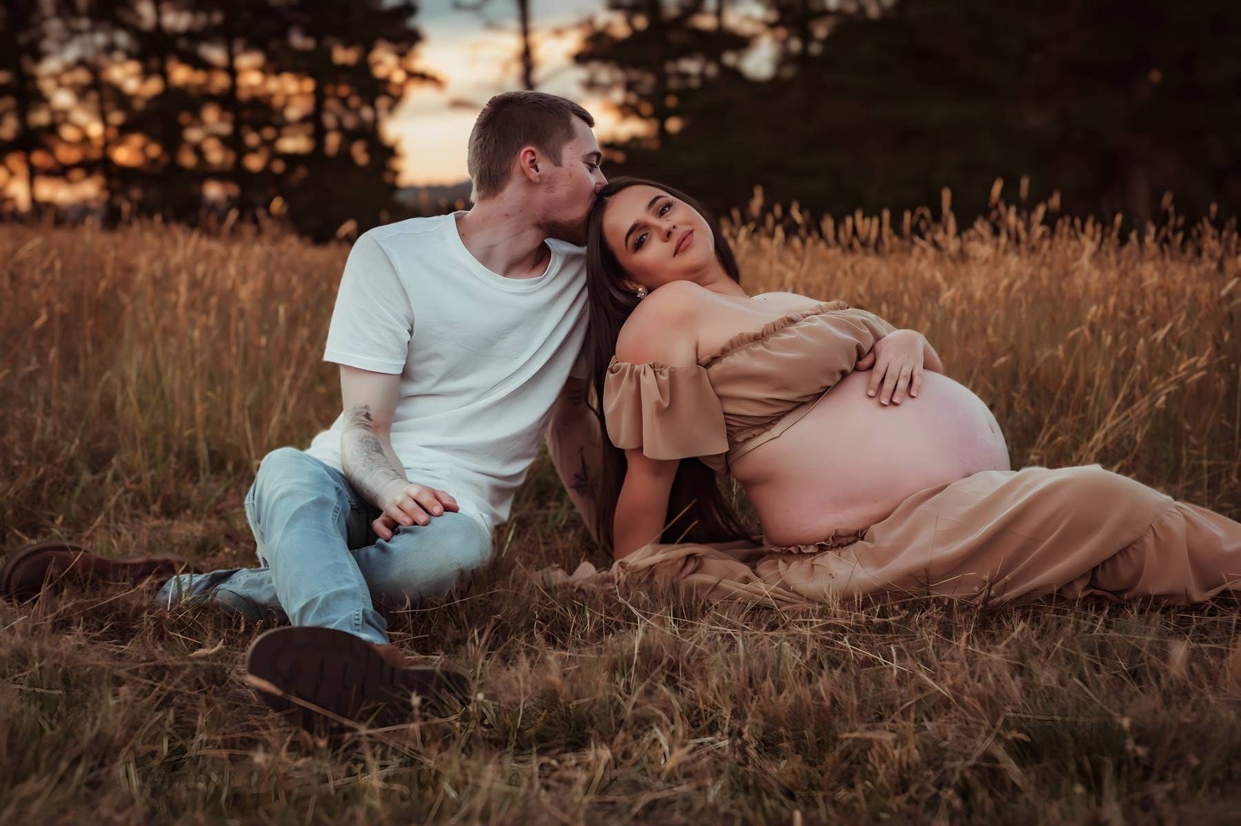 Couple sitting in golden field at sunset, pregnant woman in beige dress cradled by partner in white tee.