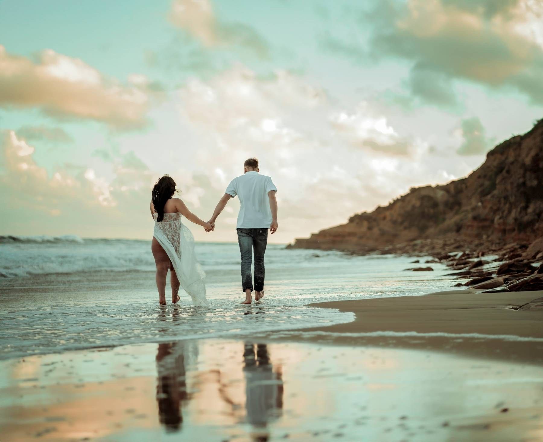 A couple walks hand-in-hand along a scenic beach at sunset, waves lapping at their feet near a rocky cliff.