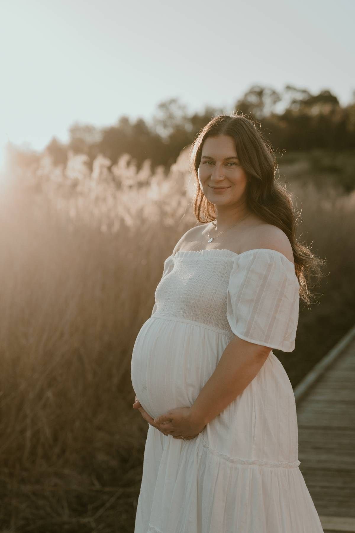 Pregnant woman in white off-shoulder dress holding baby bump at golden hour on a wooden boardwalk.