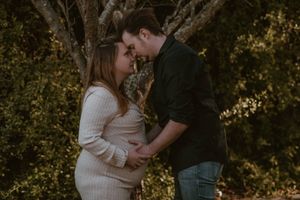 Couple sharing a tender moment outdoors, woman in white maternity dress, man in dark shirt, surrounded by lush greenery.