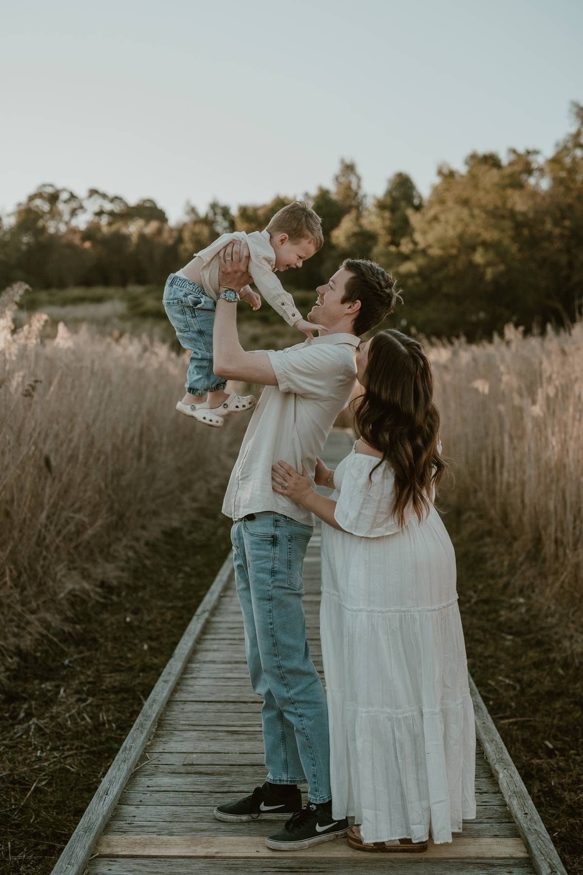 A family poses on a wooden boardwalk, father lifting toddler while pregnant mother watches, surrounded by golden fields.