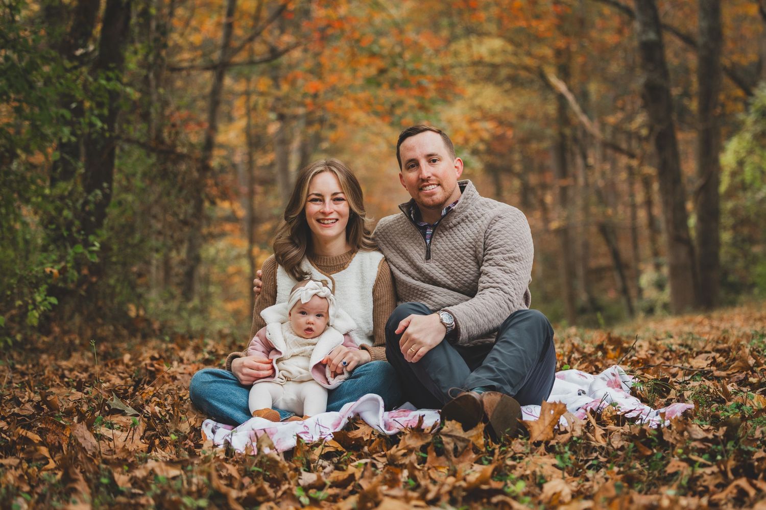 Family of three sitting on autumn leaves in a forest, smiling couple with baby in white outfit during fall foliage season.