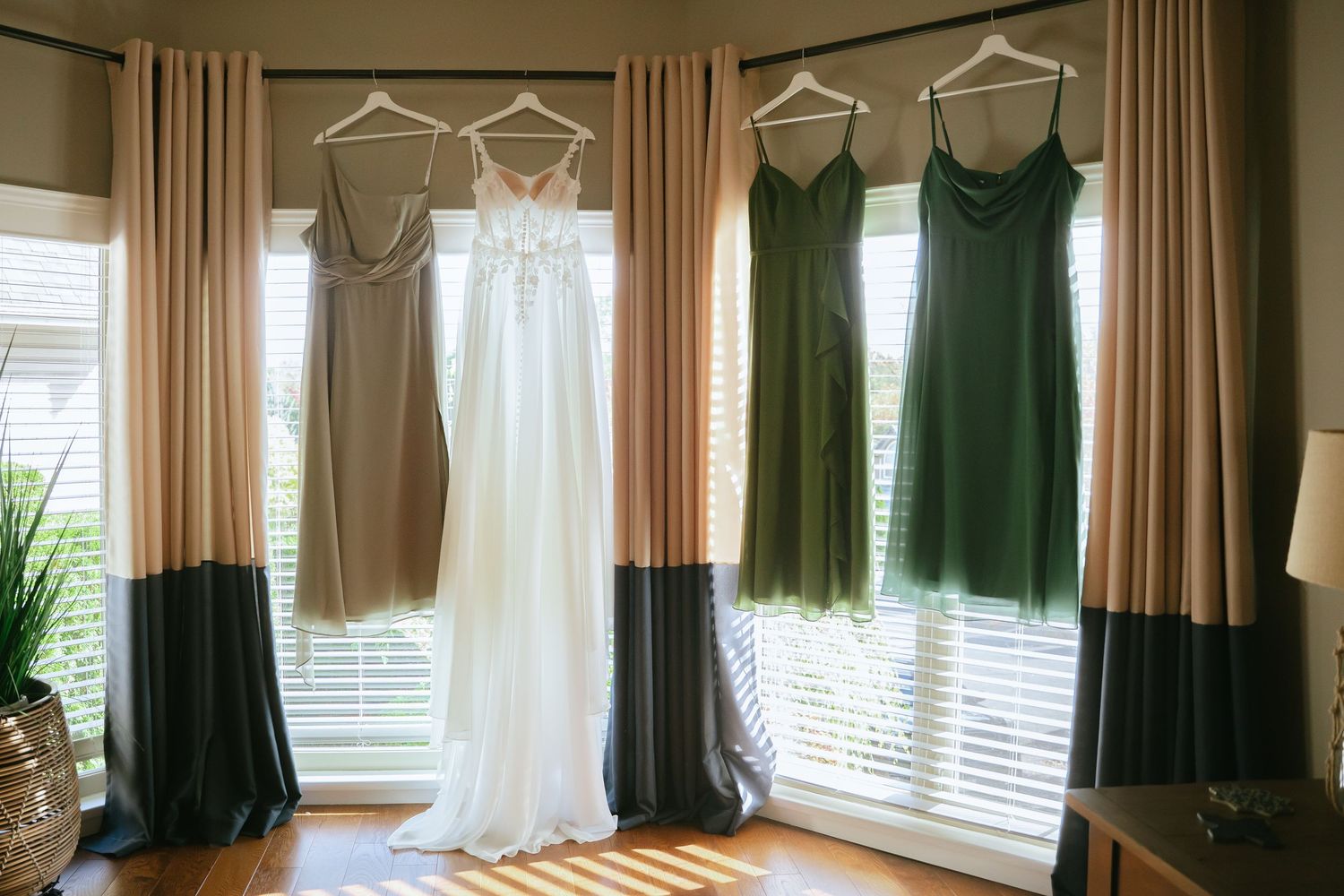 Melissa's white wedding dress hanging beside green bridesmaid dresses in front of bright windows.