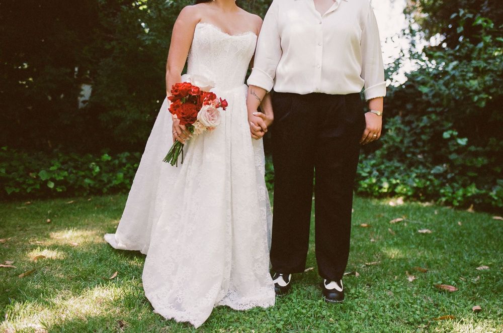 Bride in a white lace wedding gown holding a red and pink bouquet while holding hands with a partner in black pants on a sunlit green lawn.