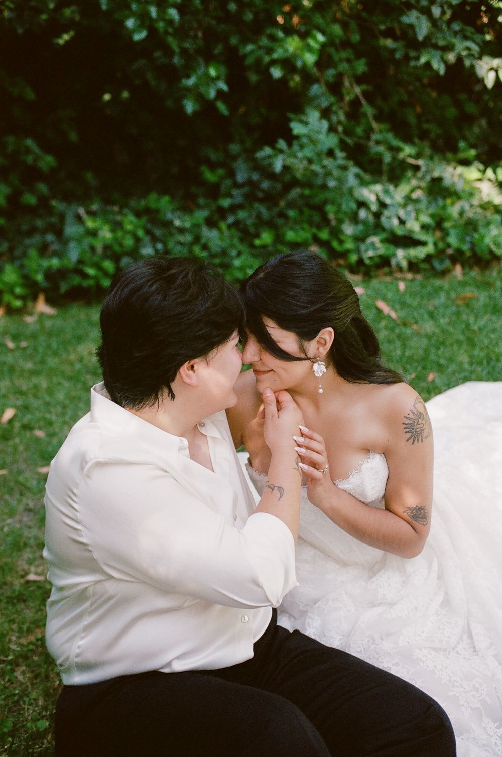 Two brides share an intimate moment on a lawn, one in a white wedding gown with tattoos and the other in a white shirt and black pants.