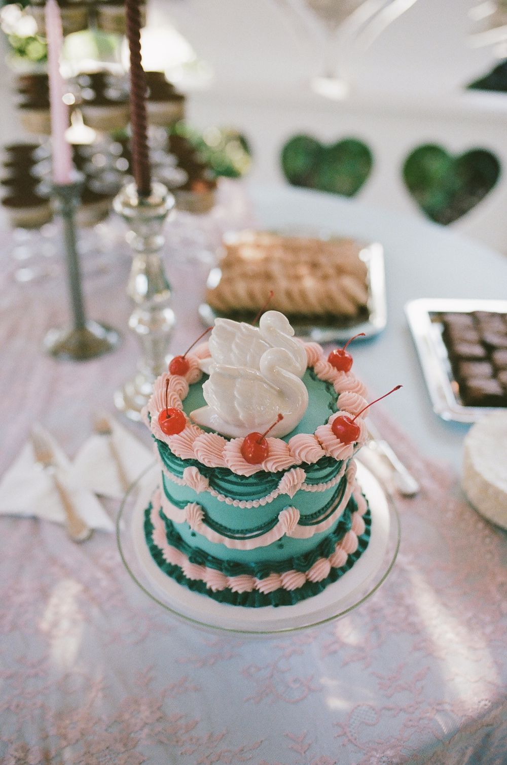 Vintage-style teal and pink frosted cake topped with a white dove and red cherries on a glass stand at a dessert table.