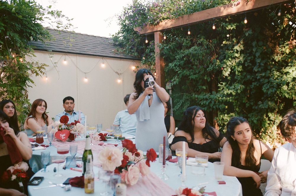 Guests seated around a long outdoor dinner table with red floral centerpieces and string lights while a woman stands taking a photo.